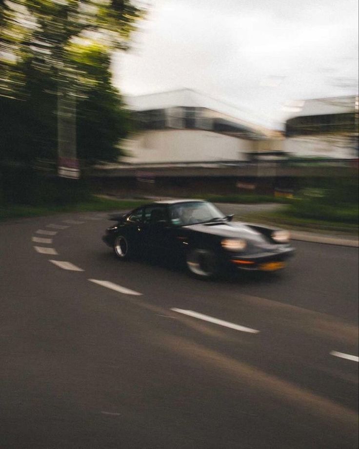 a black sports car driving down the road with trees in the background and buildings on either side