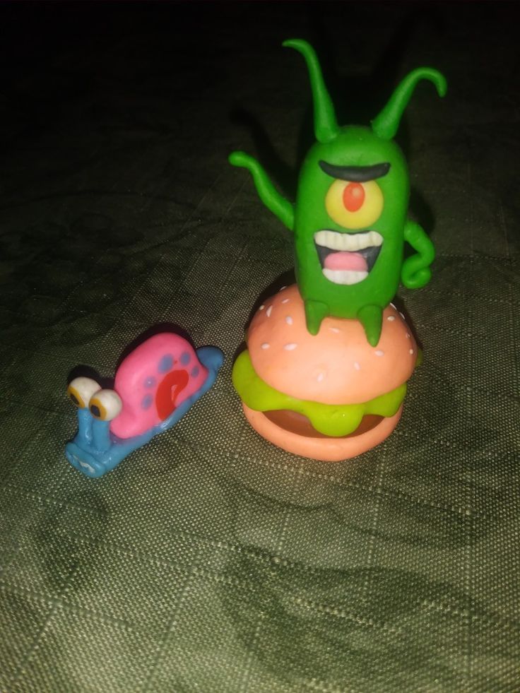 a green toy monster sitting next to a pink toy snail on top of a bed