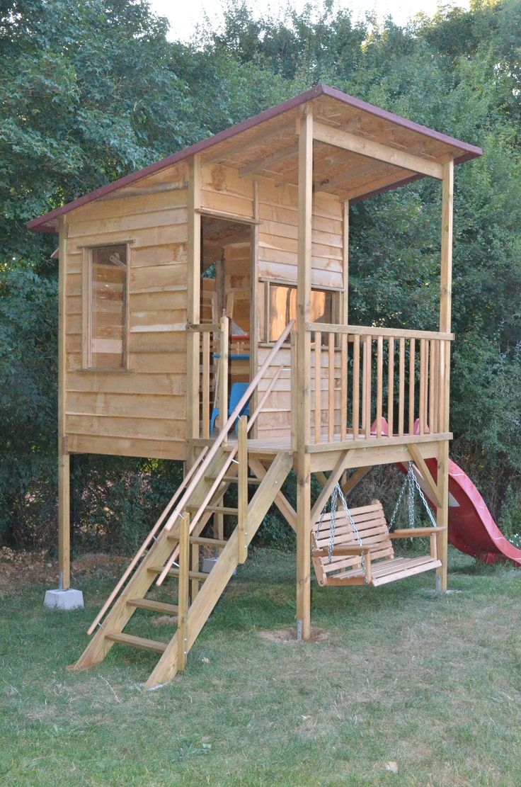 a small wooden play house with a slide and swing set in the grass next to trees