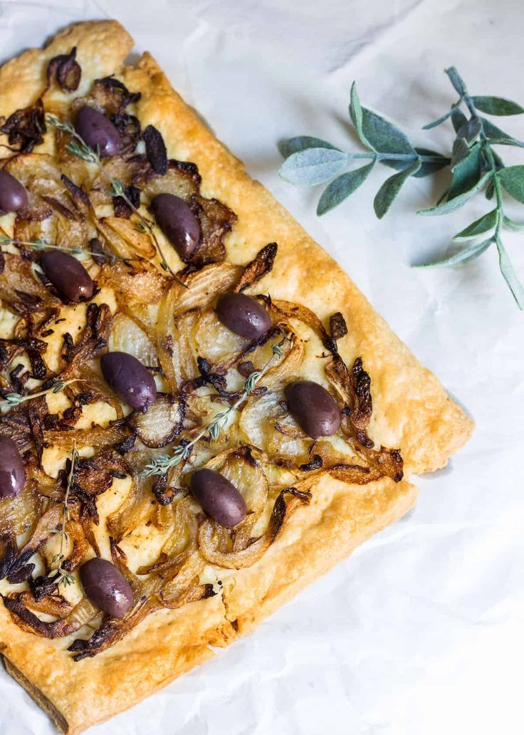 a square pizza with olives and onions on it next to a sprig of green leaves