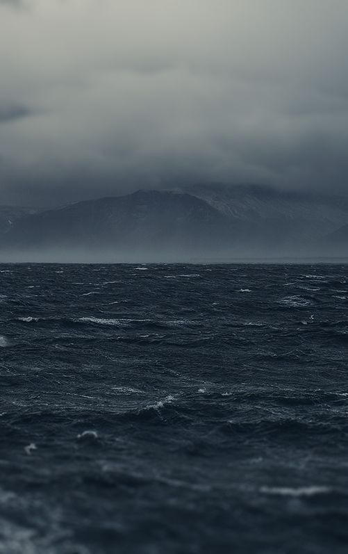 an ocean with mountains in the distance under a cloudy sky and dark clouds above it