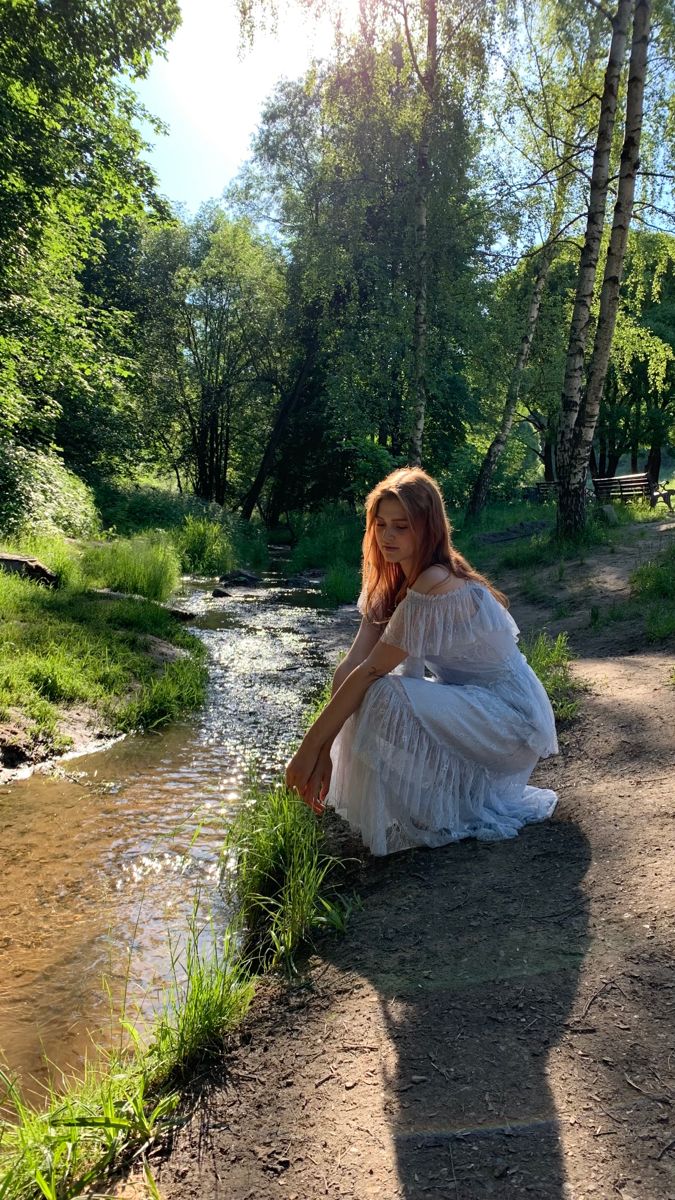 a woman in a white dress sitting on the ground next to a river and trees