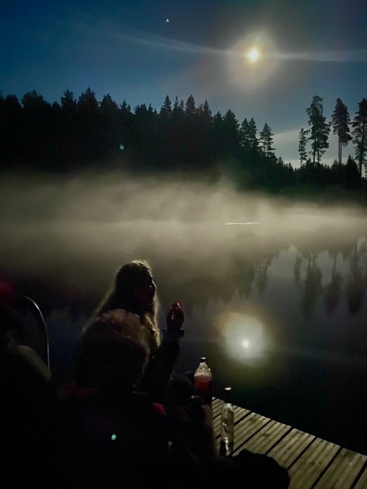 two people sitting on a dock at night with the moon in the sky above them