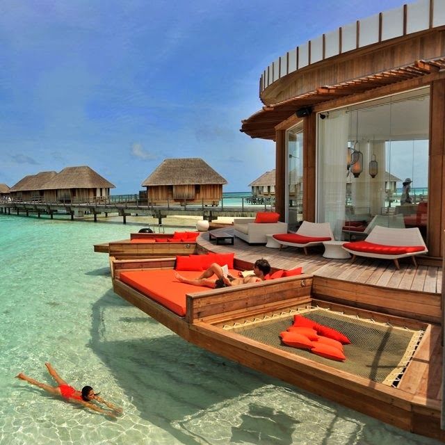 people are lounging in the water near lounge chairs and thatched huts on the beach