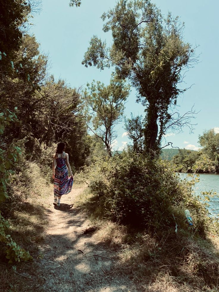 a woman walking down a dirt road next to a body of water with trees in the background