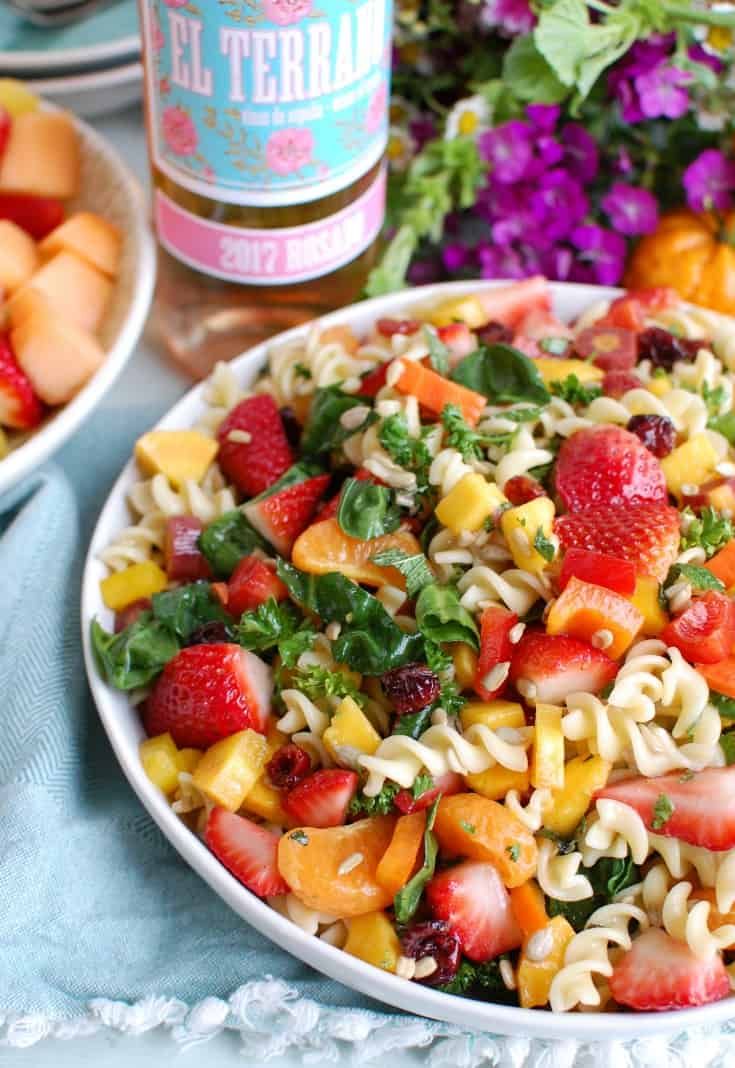 a bowl of pasta salad with strawberries and spinach next to a bottle of beer