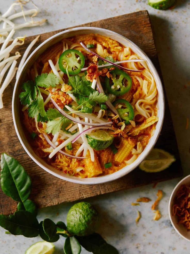 a bowl filled with noodles and vegetables on top of a wooden cutting board next to limes