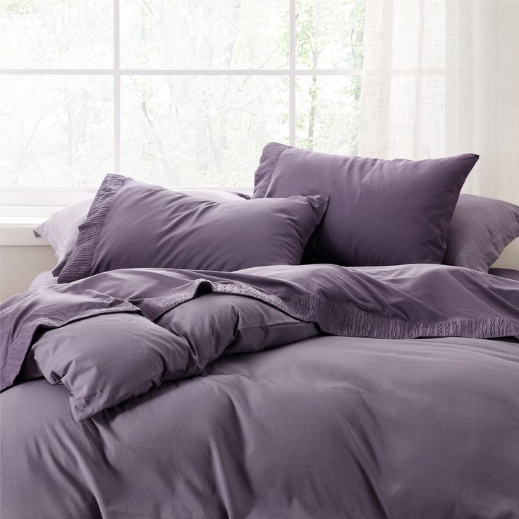 an unmade bed in front of a window with purple sheets and pillows on it