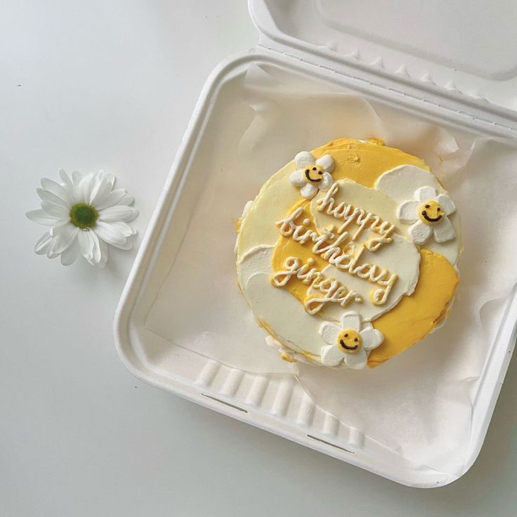 a birthday cake sitting on top of a plastic container next to a flower and a white daisy