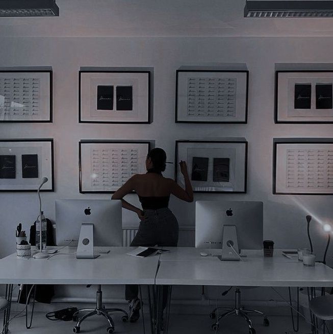 a woman sitting at a desk with two computers in front of her and several framed pictures on the wall behind her