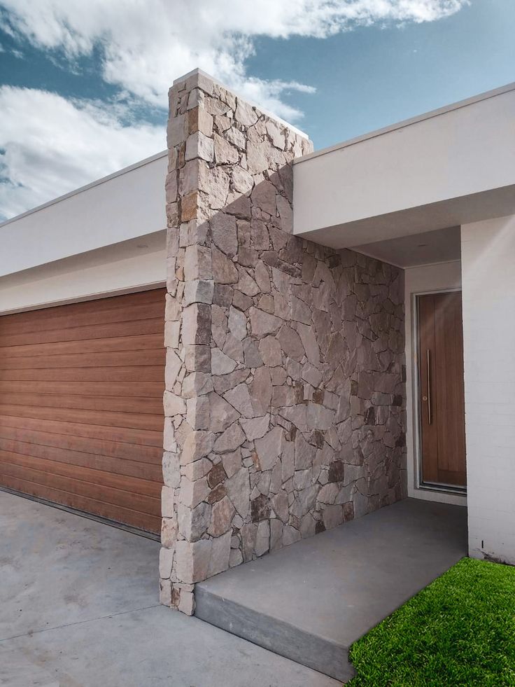 a house with a large stone wall next to a garage door and grass in front of it