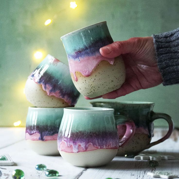 three coffee mugs that have been painted with different colors and shapes, being held by someone's hand