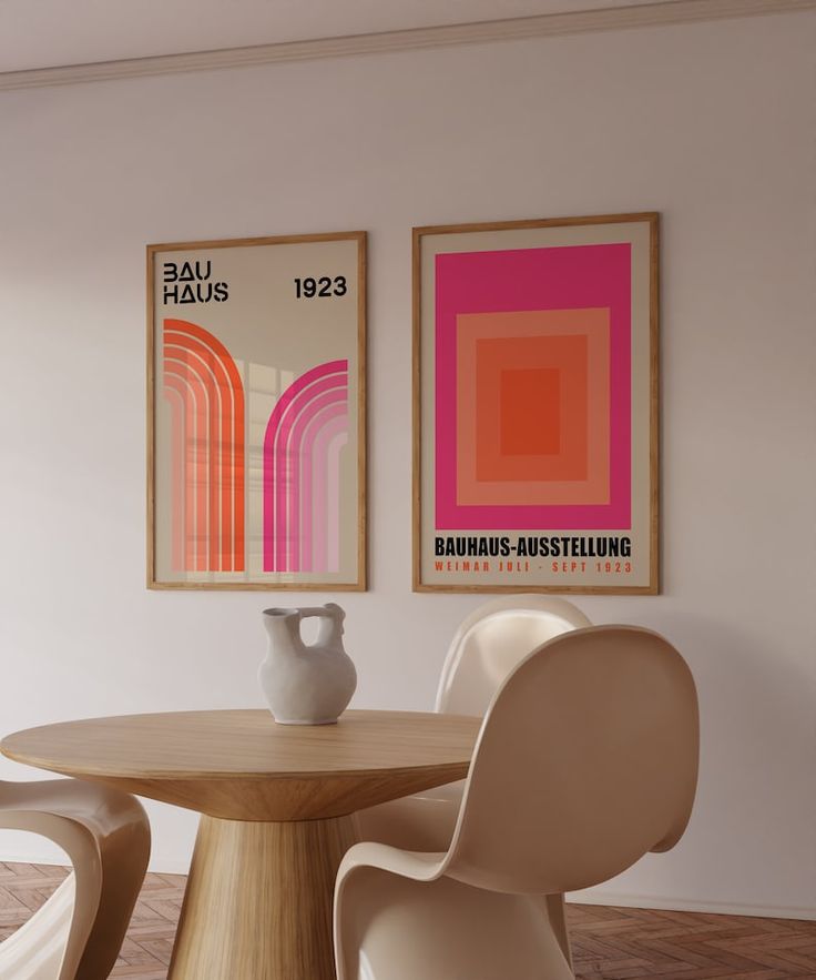 two paintings hang on the wall next to a round table with white chairs and a vase