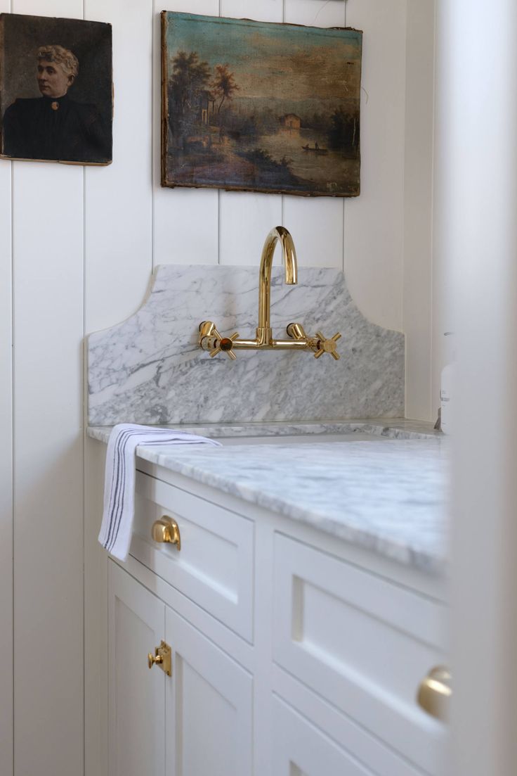 a marble sink with gold faucets in a white kitchen area next to paintings on the wall