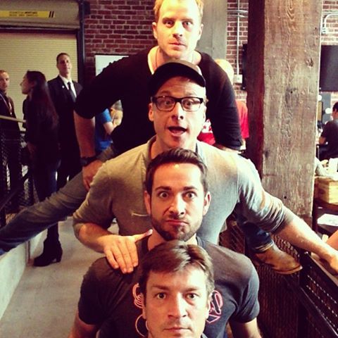 four men are posing for the camera in front of a brick wall with people standing around