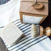 a wooden shaving brush next to a soap bar on a striped towel with a brown box