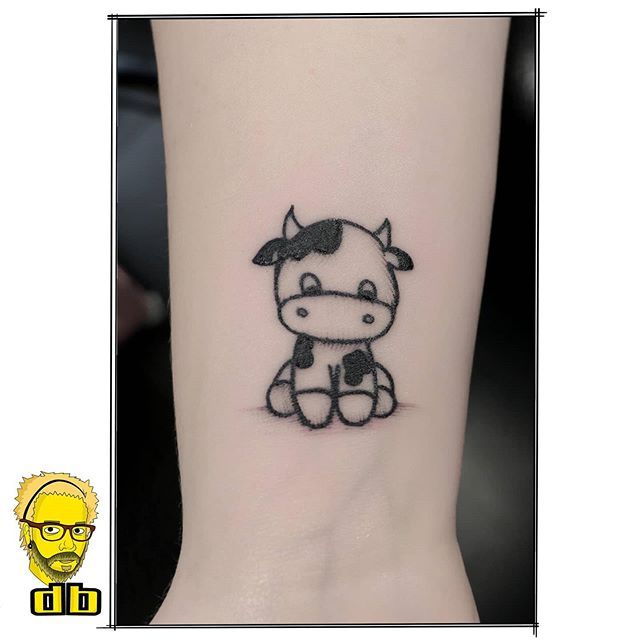 a small black and white cow tattoo on the ankle, next to a cartoon character