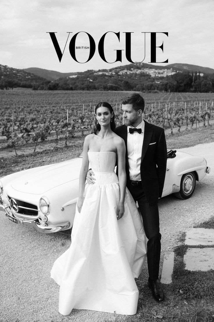 a bride and groom standing in front of a classic car on the road near a vineyard