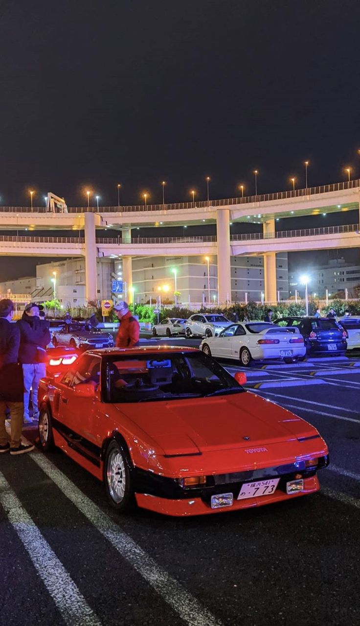 two people standing next to a red sports car in a parking lot with other cars