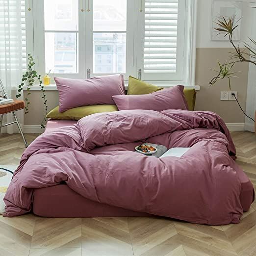 a bed with pink comforter and pillows in a room next to two large windows