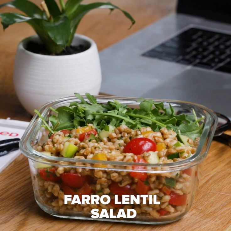 a salad in a glass container on a wooden table next to a laptop and potted plant