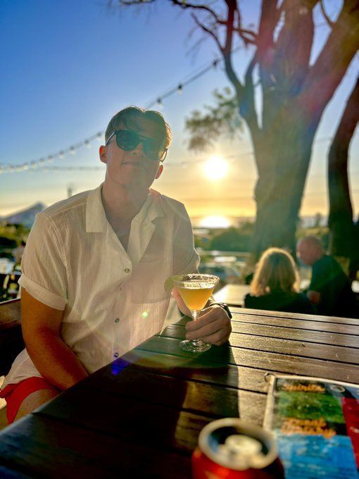 a man sitting at a table with a drink in front of him and the sun behind him