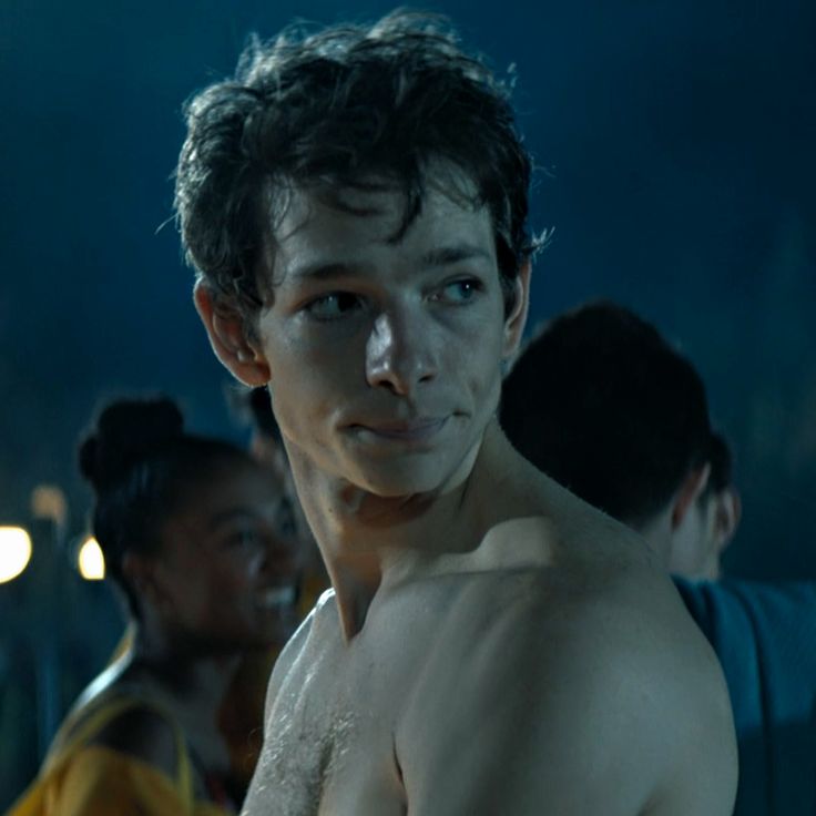 a shirtless young man standing in front of other people at night time, with one hand on his chest