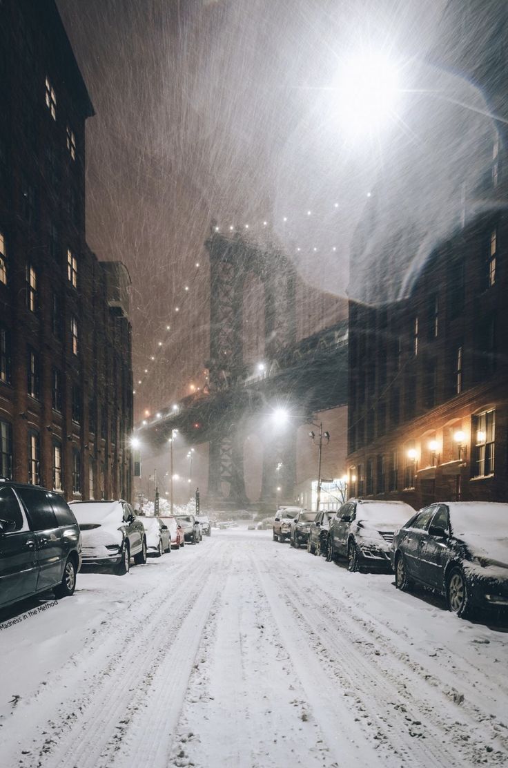 cars are parked in the snow on a city street at night with buildings and lights