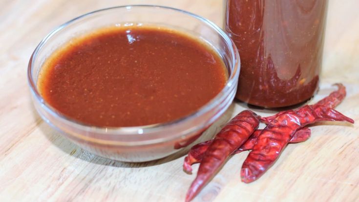 two hot sauces sit next to each other on a wooden table with red chili peppers