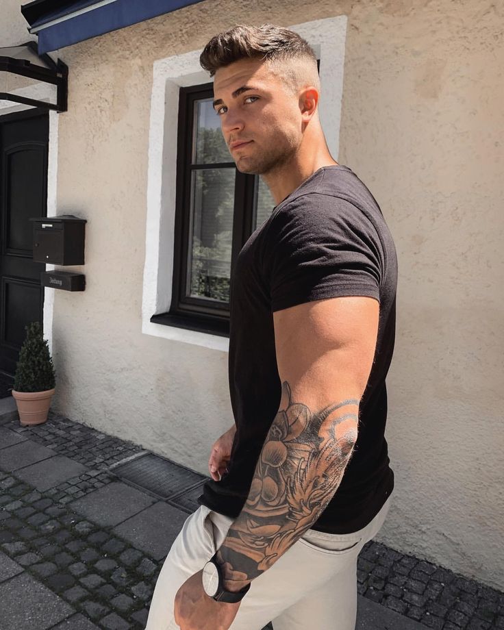 a man with tattoos standing in front of a building and looking at the camera while wearing white pants