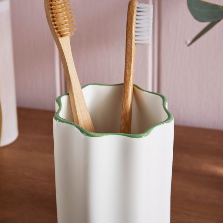 two wooden toothbrushes in a white cup on a table next to a plant