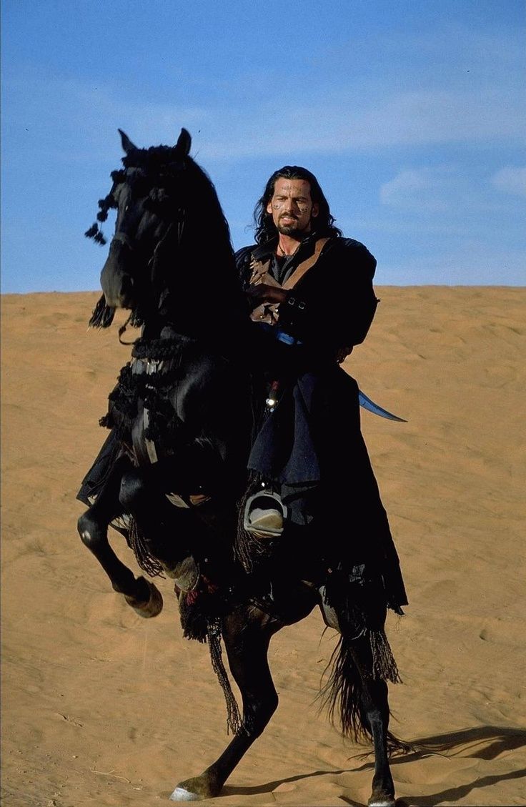 a man riding on the back of a black horse in the middle of a desert