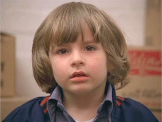 a young boy with blonde hair and blue jacket looking at the camera while standing in front of boxes