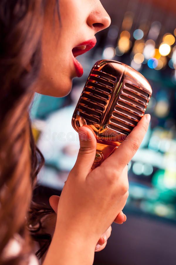 a woman singing into a microphone in front of a bar with lights on the background