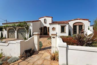 a white stucco house with red tile roofing and door, surrounded by greenery