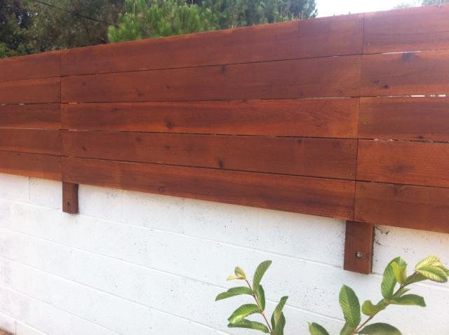 a wooden fence is next to a white brick wall with green plants in the foreground