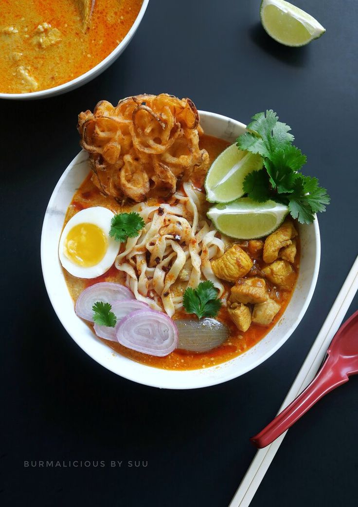 a bowl filled with noodles, meat and vegetables next to a spoon on a table