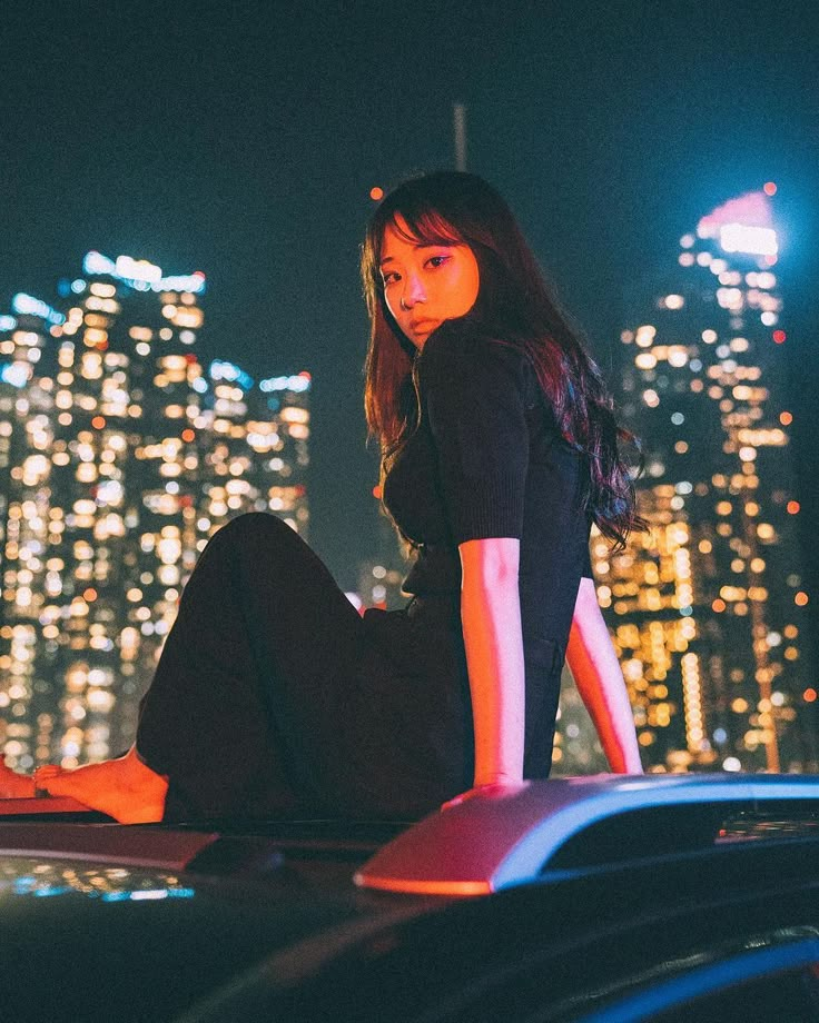 a woman sitting on the hood of a car in front of a city at night