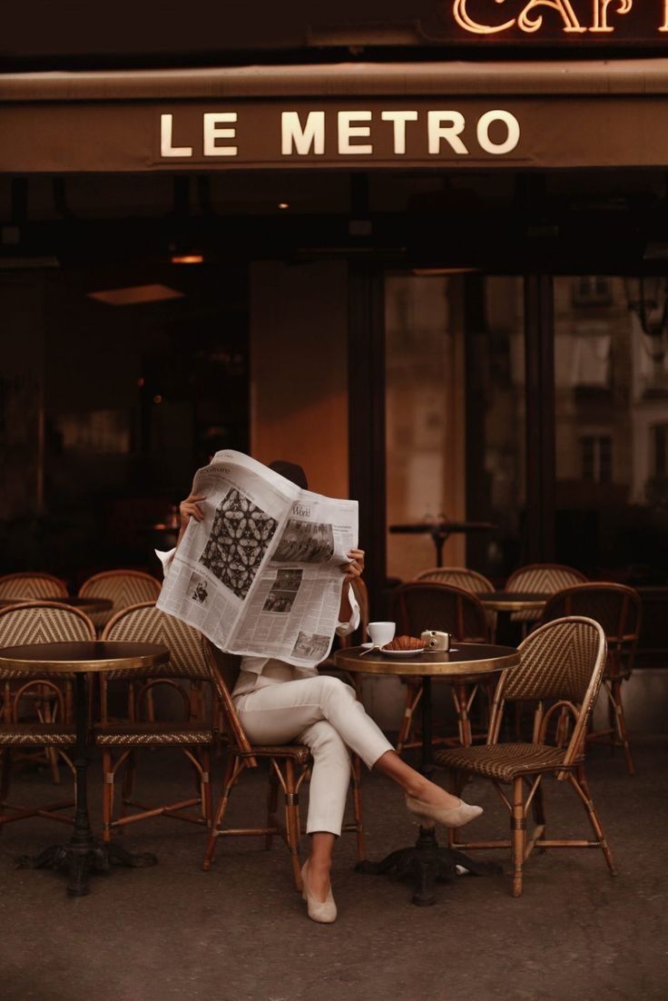 a woman sitting at a table in front of a cafe reading a newspaper with her legs crossed