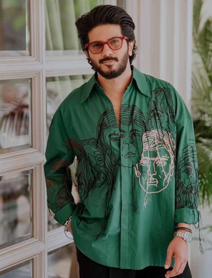 a man with glasses standing in front of a window wearing a green shirt and black pants