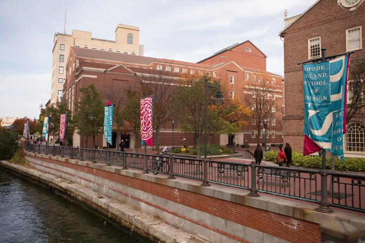 people are walking on the side of a river near buildings and banners in front of them