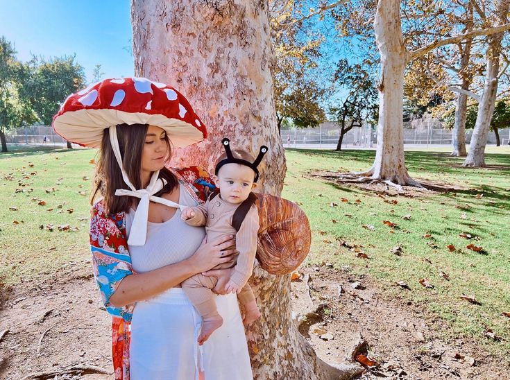 a woman holding a baby in her arms while standing next to a tree with a mushroom hat on