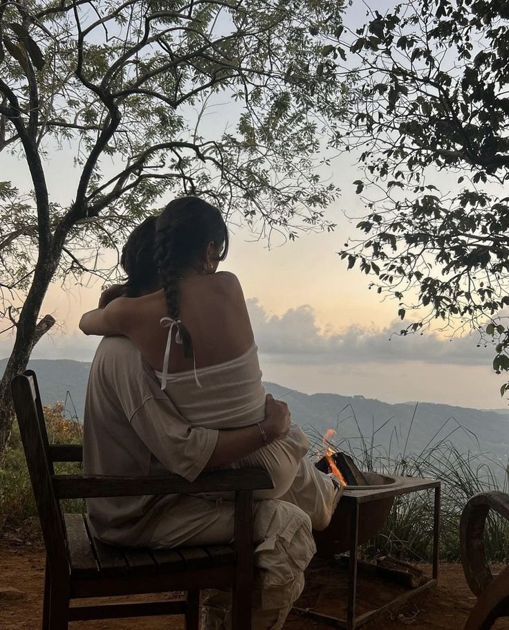 a woman sitting on top of a wooden chair next to a fire in the forest