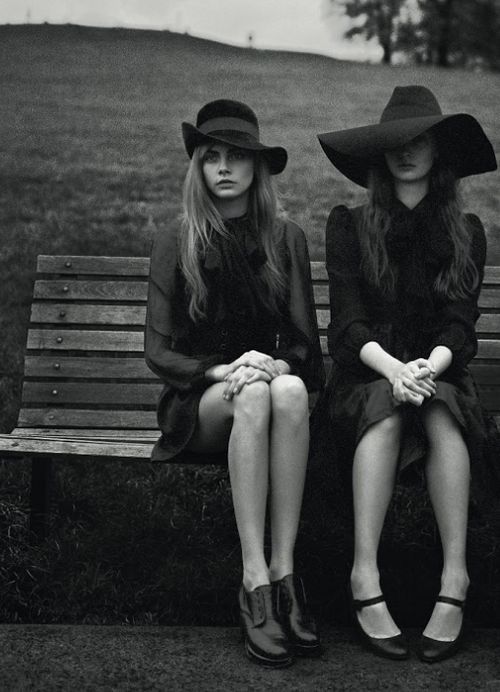 two women sitting on a park bench wearing hats and long black dresses with their legs crossed