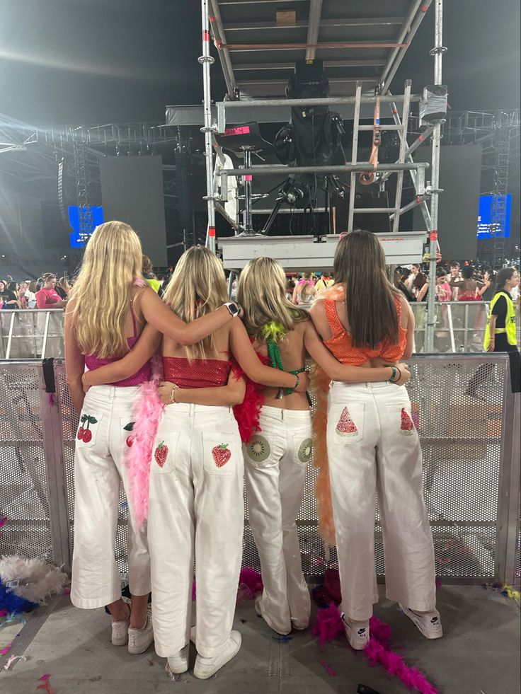 four girls in white pants and strawberries on their backs, standing together at a concert