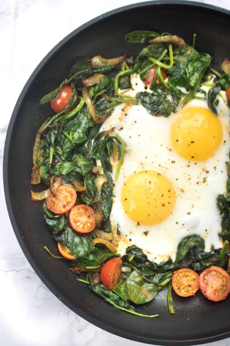 two fried eggs on top of spinach in a skillet with tomatoes and peppers