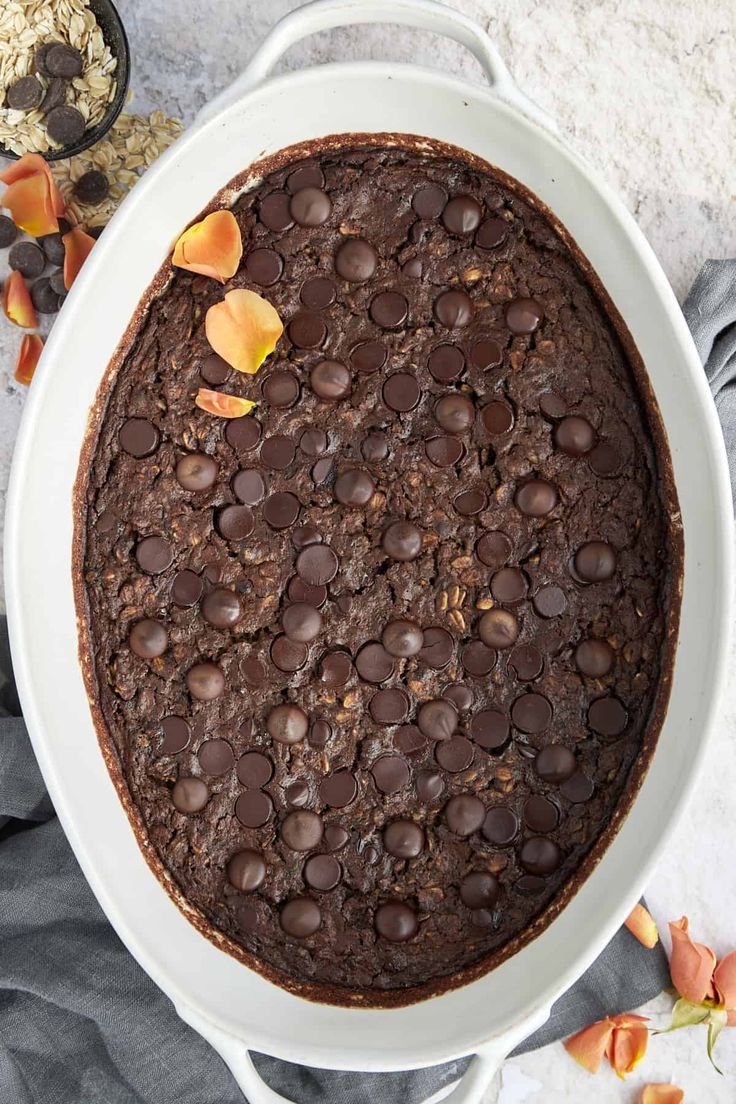 a chocolate cake in a white dish on top of a gray cloth with flowers around it