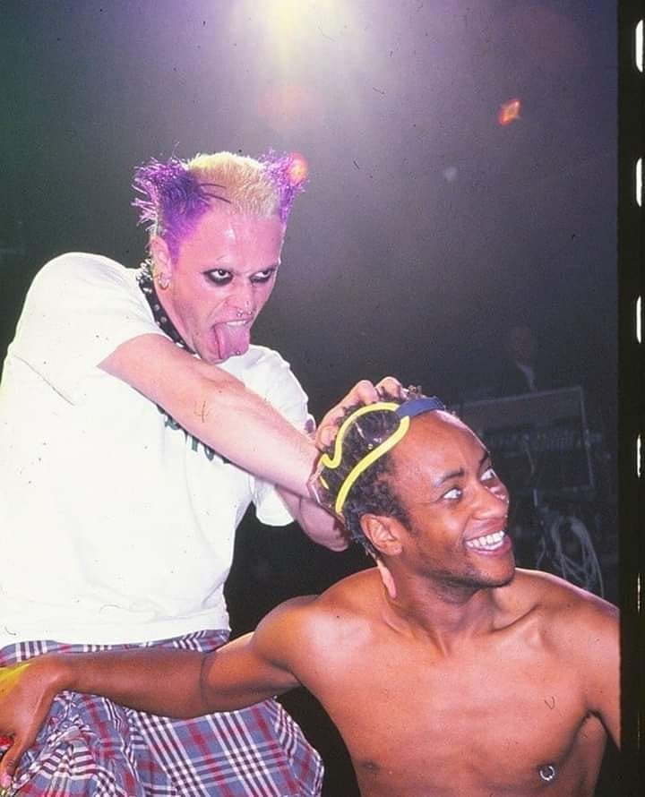 a man is cutting another mans hair with a purple mohawk on his head and wearing plaid shorts