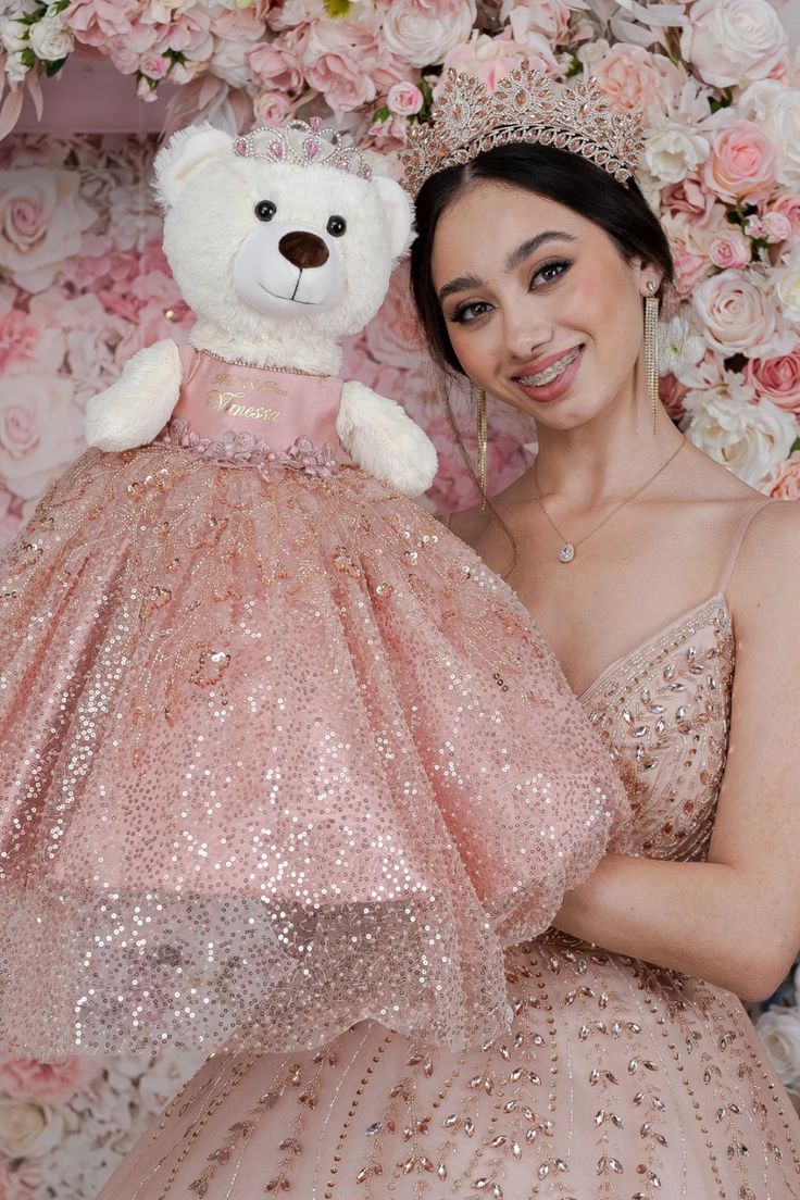 a woman in a pink dress holding a teddy bear and posing for the camera with flowers behind her