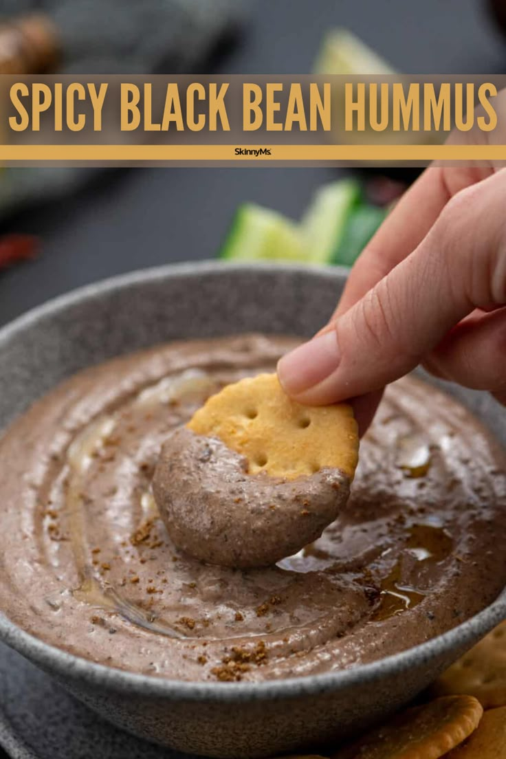 a hand dipping a cracker into a bowl of hummus with the words spicy black bean hummus above it